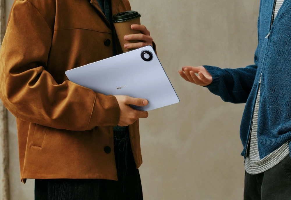 Kahverengi ceketli bir kişi sol elinde arkası dönük ve kamera modülü görünen açık renkli bir Huawei MatePad tablet tutarken, sağ elindeki kahve bardağıyla yanında duran, uzanmış eliyle iletişim kuran diğer öğrenci veya profesyonel bireyle bir araya geliyor.