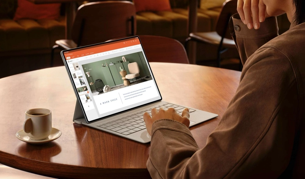 Yuvarlak ahşap bir masada oturan, sırtı dönük bir kişinin kahverengi ceket giymiş haliyle Huawei MatePad Pro Max tableti klavye eklentisiyle kullanarak iç mimari sunumu üzerinde çalıştığı görülüyor; bu düzenek, hem öğrencilerin hem de profesyonellerin yoğun iş akışlarında tablet kullanımını yansıtıyor, yanında bir fincan kahve de duruyor.