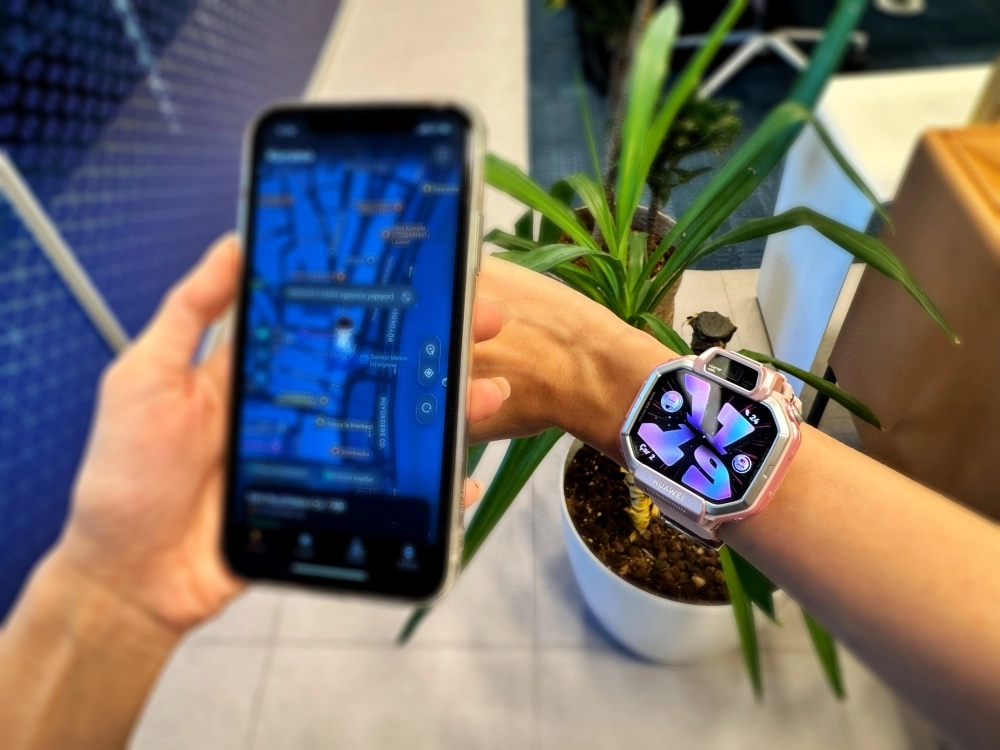 Bir eliyle bulanık bir konum takip haritası gösteren akıllı telefonu tutan ve diğer bileğinde pembe-gümüş renkli Huawei Watch Kids X1 akıllı saati takılı olan bir kişi, cihazın konum takibi özelliğinin kullanımını gözler önüne seriyor.