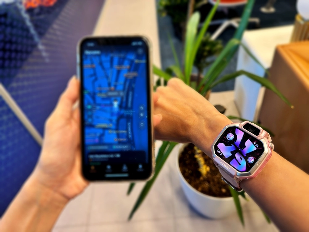 Elinde harita uygulaması açık bir akıllı telefon tutan kişinin sağ bileğinde, çocukların konumunu takip etme özelliğini gösteren renkli ekranlı pembe bir Huawei Watch Kids X1 akıllı saat bulunmaktadır.