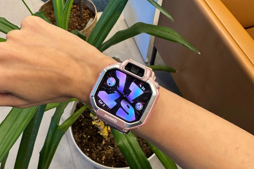 Bir kişinin bileğinde pembe renkli, üst kısmında kamerası bulunan ve parlak mor-mavi tonlarında soyut bir arayüz gösteren bir Huawei çocuk akıllı saati yakın çekimde görünüyor; arka planda yeşil yapraklı bir bitki ve kahverengi bir koltuk seçilebiliyor.