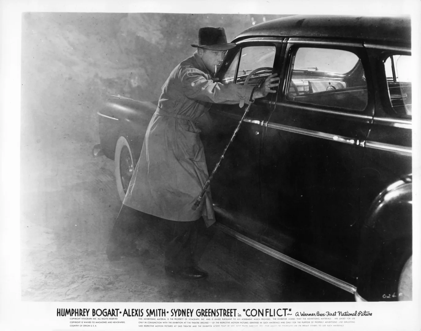 İkonik bir aktör olan Humphrey Bogart, bir film karesinde fedora şapkası ve bej trençkotuyla karanlık, dumanlı bir arka planda eski bir arabanın kapısına yaslanmış halde elinde bastonu ve ağzında sigarasıyla şehir stilinin zamansız duruşunu sergiliyor.