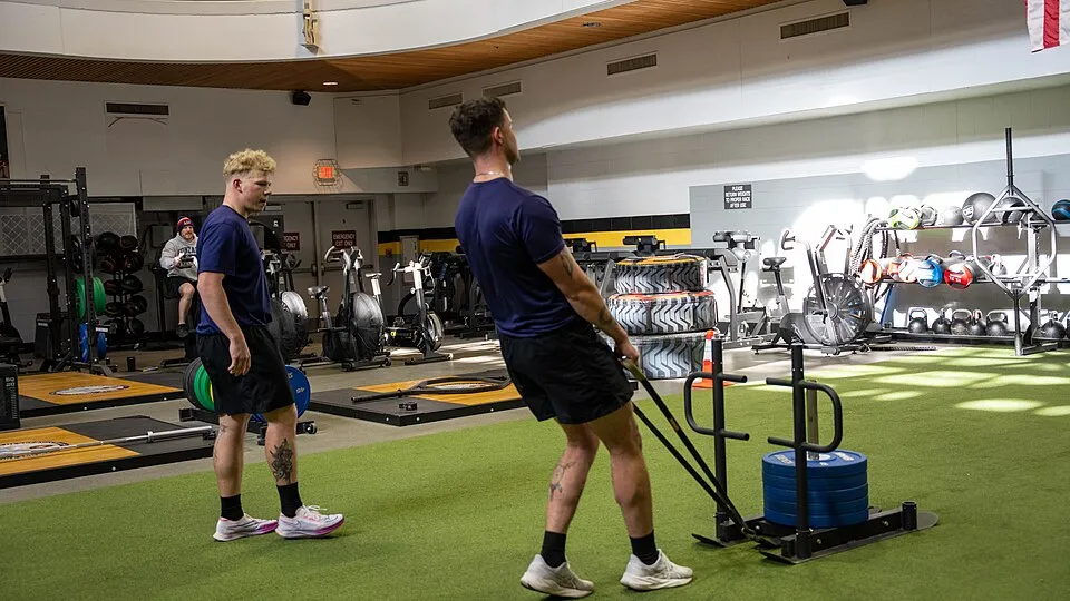 İki sporcu, yeşil suni çim kaplı geniş bir salonda, bir yandan ağır bir kızak çekme egzersizi yaparken, diğer yandan çeviklik ve kuvvet odaklı HYROX antrenmanının gerektirdiği diğer ekipmanların da bulunduğu bir ortamda çalışıyor.