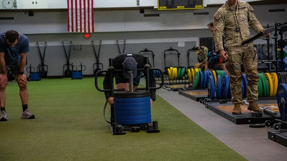 Yeşil zeminli bir spor salonunda, siyah şapkalı bir sporcu Hyrox yarışmalarında görülen ağırlıklı kızak itme istasyonunda mavi disklerle dolu kızağı iterken, solunda mola veren başka bir sporcu ve sağında askeri üniformalı bir eğitmen duruyor.