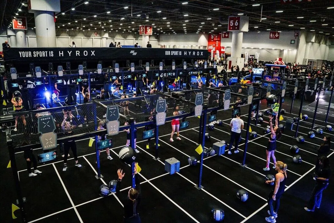 YOUR SPORT IS HYROX yazılı dev panoların altında, İstanbul'daki geniş bir kapalı spor salonunda düzenlenen Hyrox yarışmasında çok sayıda sporcu; sağlık topu fırlatma gibi çeşitli fonksiyonel fitness istasyonlarında yoğun bir şekilde antrenman yaparken görülüyor, her bir katılımcı kendi parkurunda performans için mücadele ediyor.