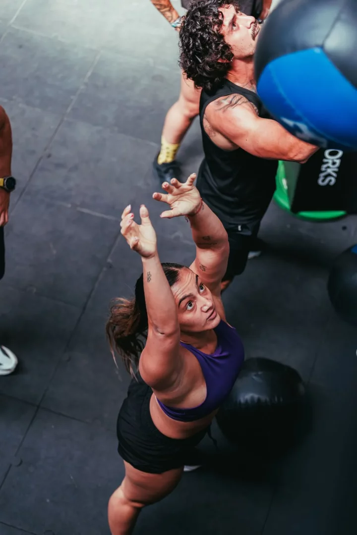 Yüksek açıdan çekilmiş bir spor salonu fotoğrafında, mor spor sütyeni ve siyah şort giyen bir kadın, kollarını yukarı uzatarak başının üzerinde bir topu yakalamak veya atmak üzereyken odak noktasıdır; arkasındaki kıvırcık saçlı erkek ise göğüs hizasında mavi-siyah bir sağlık topunu tutarak Hyrox antrenmanlarının dinamik ve fonksiyonel doğasını yansıtan bir egzersiz anını sergiliyor.