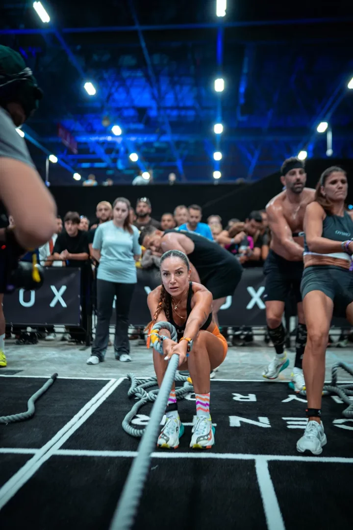 Hyrox yarışmasında Selin Sargut, dikkatini tamamen verdiği bir anla, siyah bir spor sütyeni ve turuncu şortuyla, kalın bir halatı iki eliyle sıkıca kavrayarak yere yakın bir pozisyonda çekiyor; arkasında mavi ışıklarla aydınlatılmış bir salonda diğer sporcular ve izleyiciler flu görünüyor.