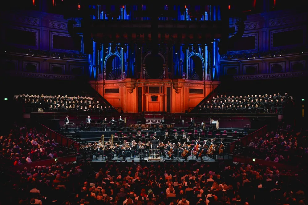 Büyük bir konser salonunda, şef yönetimindeki Royal Philharmonic Orchestra, orkestra üyeleri ve arkalarındaki geniş koro ile sahnede izleyicilere bir konser sunarken, salonun görkemli mimarisi turuncu ve mavi ışıklarla aydınlanıyor.
