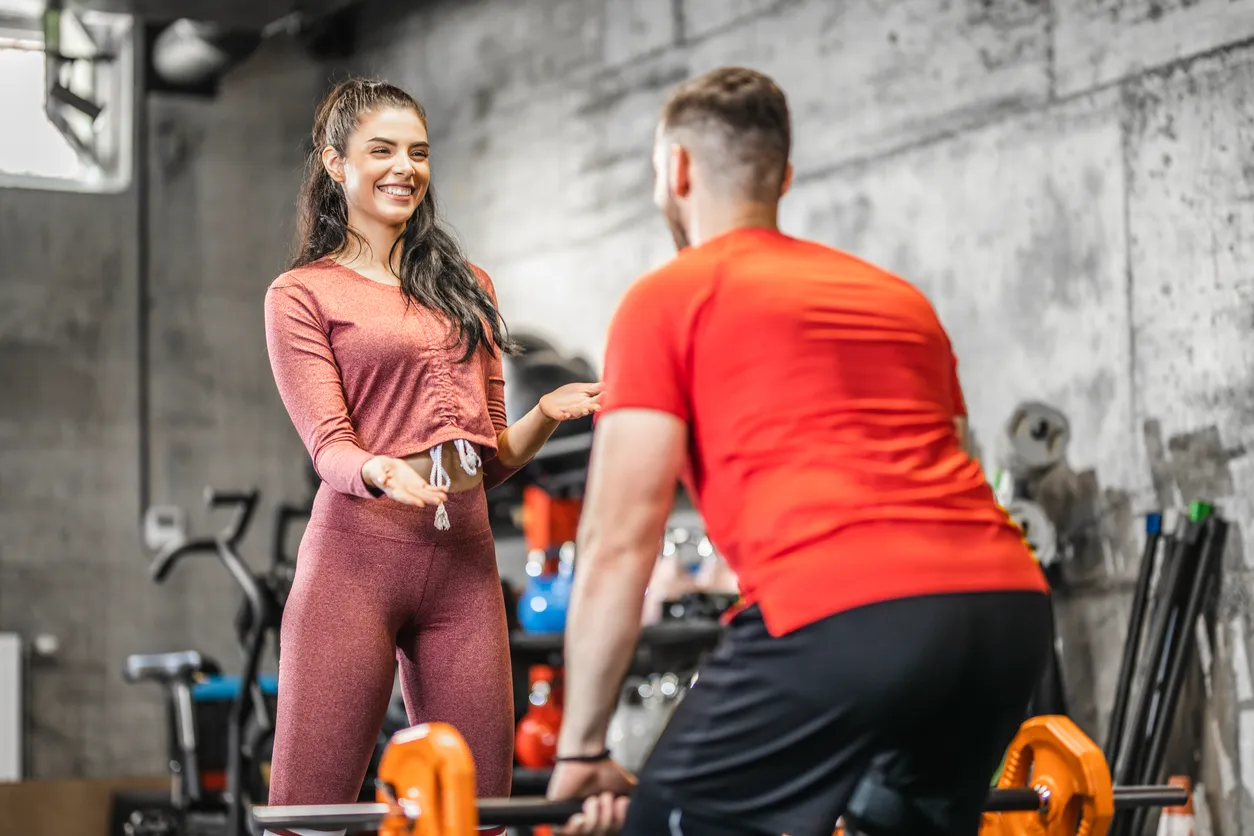 Spor salonunda, gülümseyen bir kadın spor eğitmeni veya antrenman arkadaşı, iş sonrası spor yapmak için halterle çalışan kırmızı tişörtlü bir erkeğe, beton duvara yaslanmış ekipmanların arasında, destekleyici jestlerle talimat veriyor.