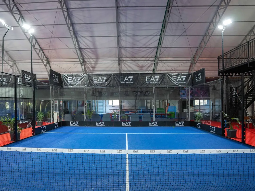 EA7 logolu panolarla süslenmiş, parlak ışıklarla aydınlatılmış, cam ve tel örgülerle çevrili, ortasında file bulunan mavi zeminli bir padel kortu, İstanbul'daki modern spor tesislerini yansıtan kapalı bir alanda görülüyor.