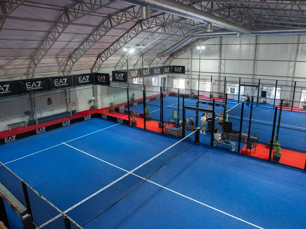 İstanbul'daki modern bir kapalı padel tesisinde, yüksek metal çatılı bir salonun altında, parlak mavi zeminli ve cam duvarlar ile tel örgülerle çevrili birden fazla padel kortu, kırmızı zeminli yürüme alanlarıyla birlikte geniş bir alanı kaplıyor, sağda ise birkaç kişi görülüyor.