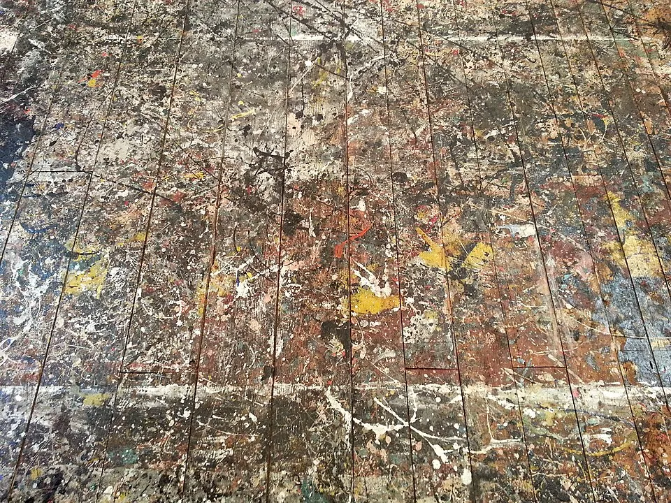 Jackson Pollock'un stüdyo zemini, sanatçının meşhur damlatma tekniğini uygularken yıllar boyunca yüzeye saçtığı siyah, beyaz, sarı, kırmızı ve mavi gibi sayısız renkteki yoğun boya sıçramalarıyla tamamen kaplı eski, yıpranmış ahşap kalasları detaylı bir şekilde gösteriyor.