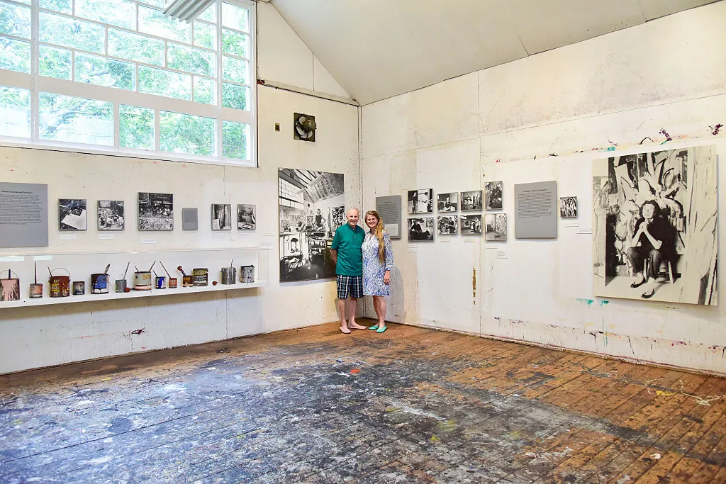 Jackson Pollock'un ünlü evi/stüdyosu olarak kullanılan ve zemini boya lekeleriyle kaplı ahşap döşemeli bu geniş odada, duvarlardaki sanatçının yaşamını yansıtan tarihi fotoğraflar ve raflardaki eskimiş boya kutuları sergisini ilgiyle inceleyen bir çift yer alıyor.