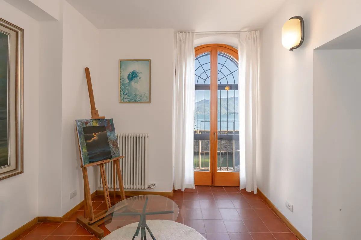 James Bond filmine ev sahipliği yapmış Villa La Gaeta'nın beyaz duvarlı ve kahverengi karolu odasında, kemerli ahşap pencereden masmavi Como Gölü ve çevreleyen dağların nefes kesen manzarası izlenirken, odanın solunda bir şövale üzerinde deniz temalı bir tablo ve sağda modern bir aplik ışığı görülüyor.