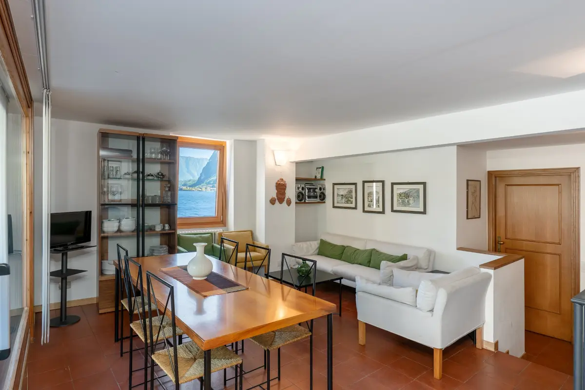 James Bond Villa La Gaeta'daki bu geniş salon ve yemek alanı, fayans zemin üzerinde hasır sandalyelerle çevrili ahşap bir yemek masası, konforlu beyaz kanepeler ve raflarında seramikler bulunan bir vitrin dolabını barındırırken, pencereden dağlarla çevrili büyüleyici bir göl manzarası odaya ferahlık katıyor.