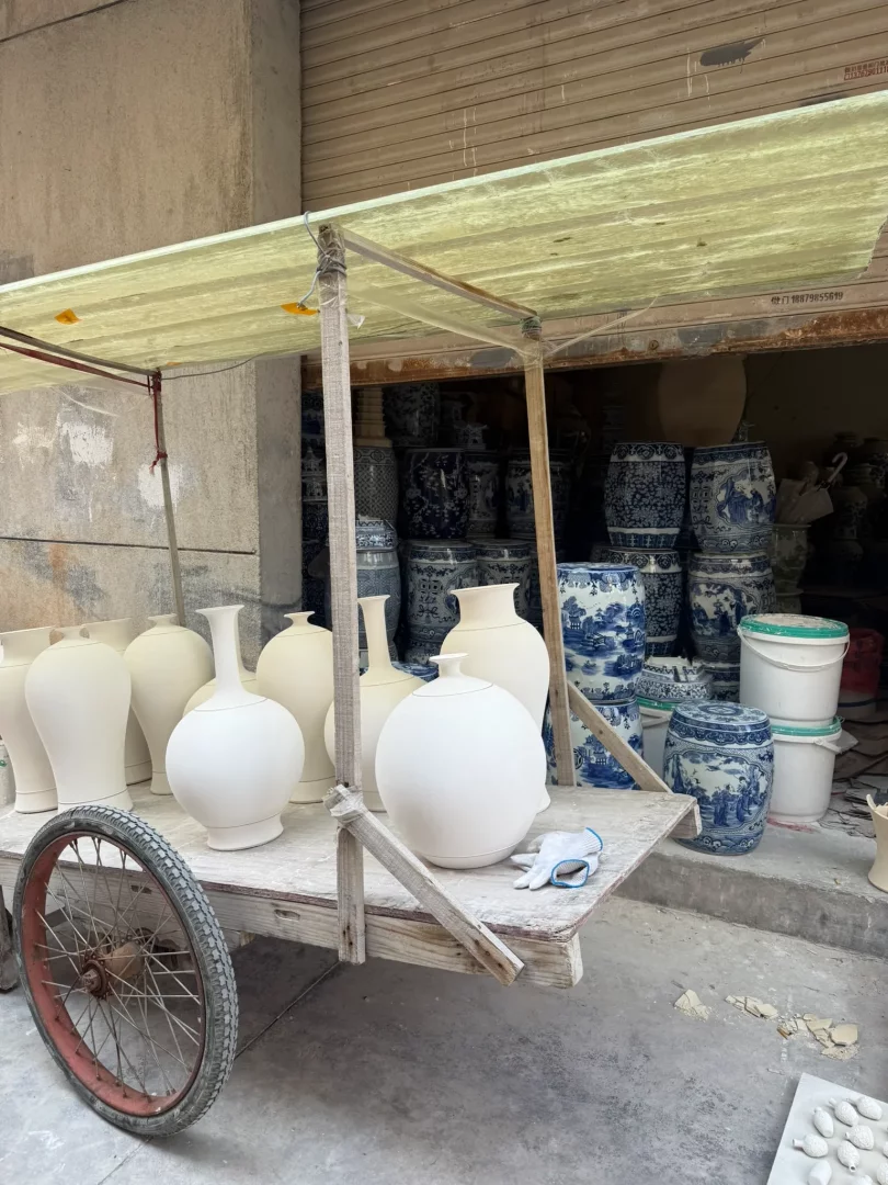 Jingdezhen'in porselen odaklı üretim kültürünün canlı bir kesitini sunan bu sokak görüntüsünde, ahşap bir el arabasının üzerinde çeşitli boyutlarda, sırsız ve porselen üretimi için bekleyen vazolar ile yuvarlak formlar dikkat çekerken, dükkanın içinde arka planda geleneksel mavi beyaz desenlere sahip, tamamlanmış porselen kaplar ve fıçılar yığılı durarak porselenin şehrin gündelik hayatının ayrılmaz bir parçası olduğunu vurguluyor.