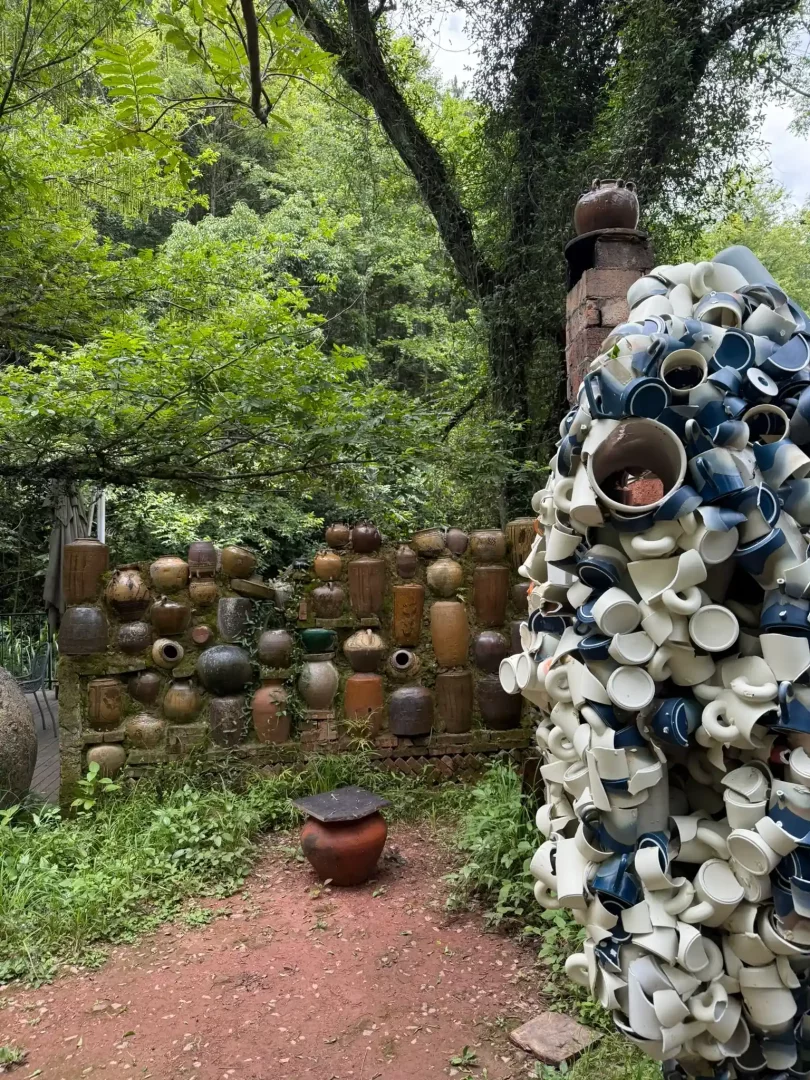 Jingdezhen'in benzersiz üretim kültürünü yansıtan bu geniş planda, yemyeşil bir bahçe içinde, yüzlerce eski porselen ve seramik kapla örülmüş bir duvar ile kırık ve sağlam fincanlardan yapılmış devasa çağdaş bir heykel porselenin burada yalnızca bir malzeme değil, hayatın kendisi olduğunu gösteriyor.