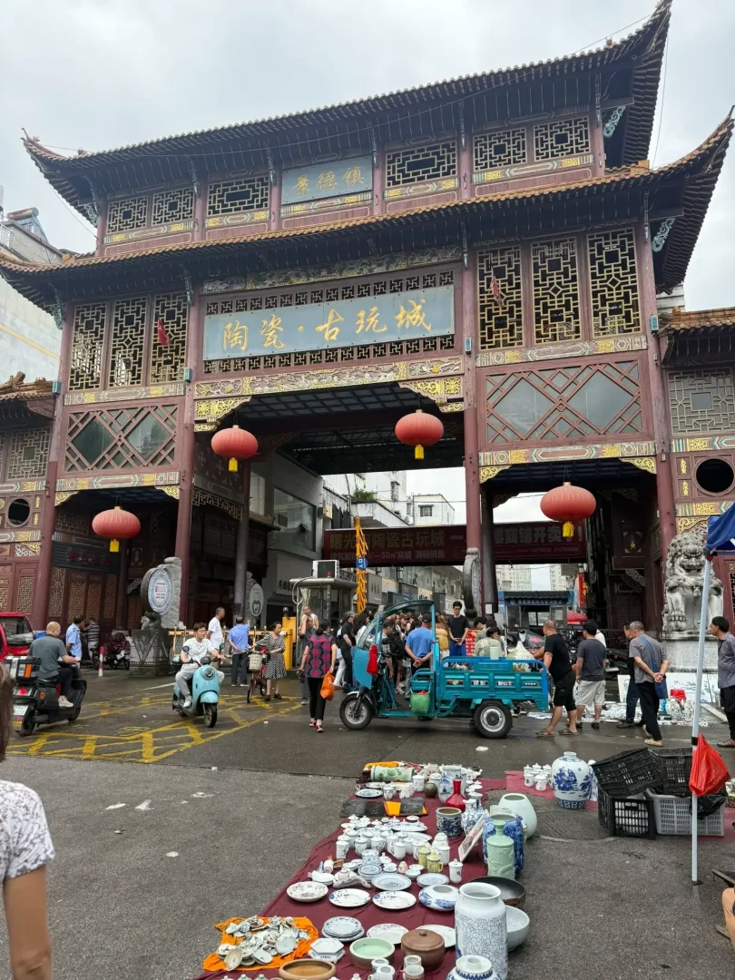 Jingdezhen'de, geleneksel Çin mimarisine sahip görkemli bir kapının önünde, günlük yaşamın ve porselen sanatının iç içe geçtiği, yerlere serilmiş antika porselenler ve çeşitli seramik ürünlerle dolu canlı bir açık hava pazarı, şehrin zengin üretim kültürünü gözler önüne seriyor.