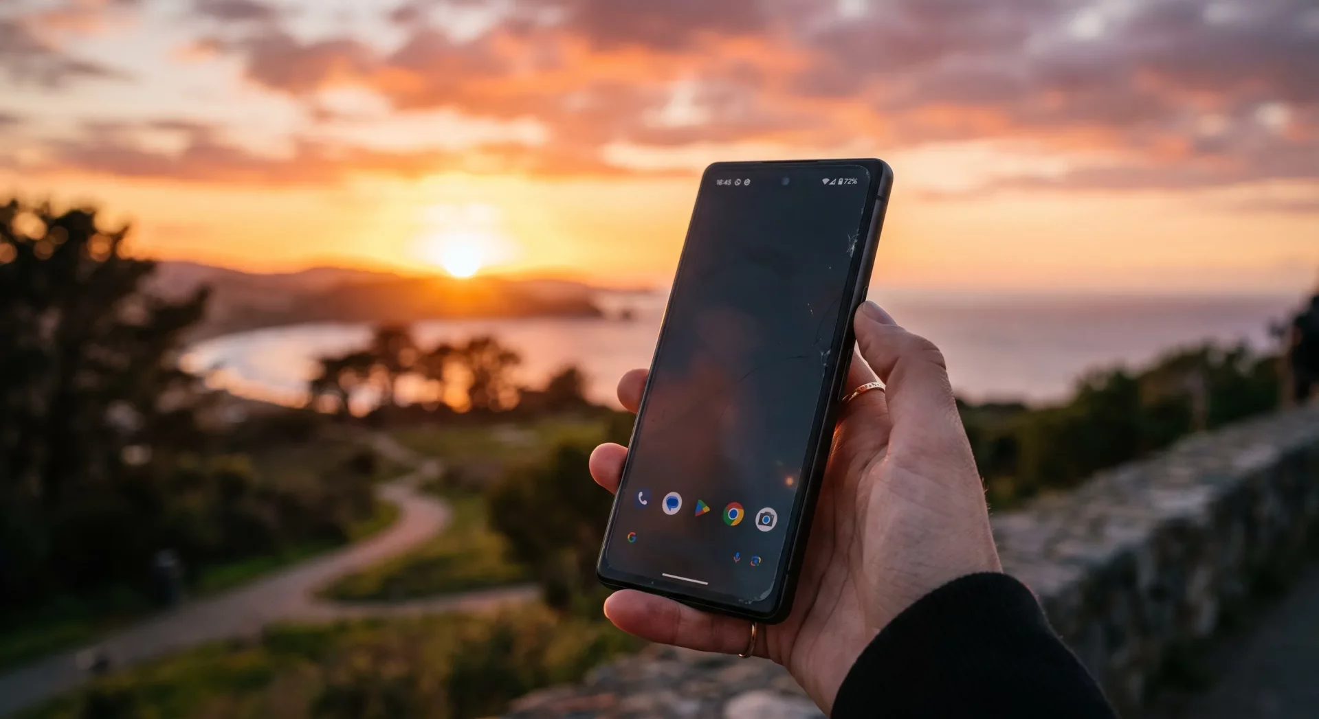 Gün batımının turuncu ve pembe tonlarıyla gökyüzünü saran, sakin bir koy manzarası önünde, çatlak ekranlı akıllı telefonunu elinde tutan bir kişi, dijital gürültüyü bilinçli olarak kısarak anı deneyimlemenin ve dış dünyayı kaçırmanın getirdiği Kaçırma Sevinci (JOMO) felsefesini simgeliyor.