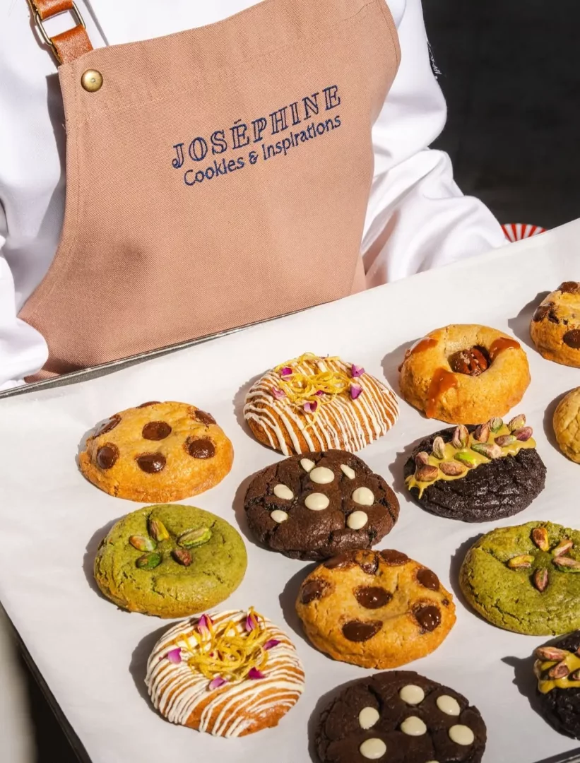 Joséphine Cookies & Inspirations yazılı bej bir önlük giyen beyaz şef önlüklü bir kişinin tuttuğu tepside, damla çikolatalı, antep fıstıklı, beyaz çikolatalı, karamelli ve limon kabuğu rendesiyle süslenmiş çok çeşitli lezzetli gurme kurabiyeler estetik bir düzenle sergileniyor.