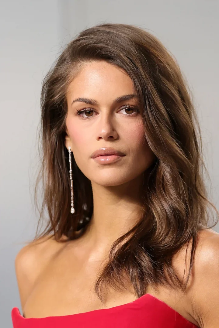 Kaia Gerber'in omuzlarından dökülen uzun, parlak kahverengi ve belirgin dalgalı saç modeli, doğal makyajı, şeffaf dudak parlatıcısı ve göz alıcı uzun pırlanta küpeleriyle kırmızı straplez kıyafetini tamamlayarak zarif bir duruş sergiliyor.