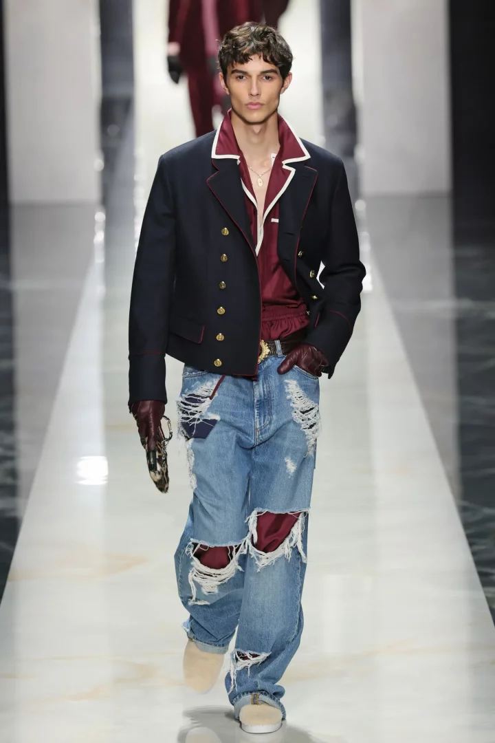 Parlak podyumda yürüyen genç erkek model, altın düğmeleri ve beyaz biyeli yakasıyla dikkat çeken lacivert kısa blazer ceketini, altına giydiği bordo gömlek ve dizleri yırtık, içinden bordo kumaş görünen açık mavi kot pantolonla kombinlemiş, ayrıca bordo deri eldivenleri ve bej makosen ayakkabıları ile doğrudan kameraya bakıyor.