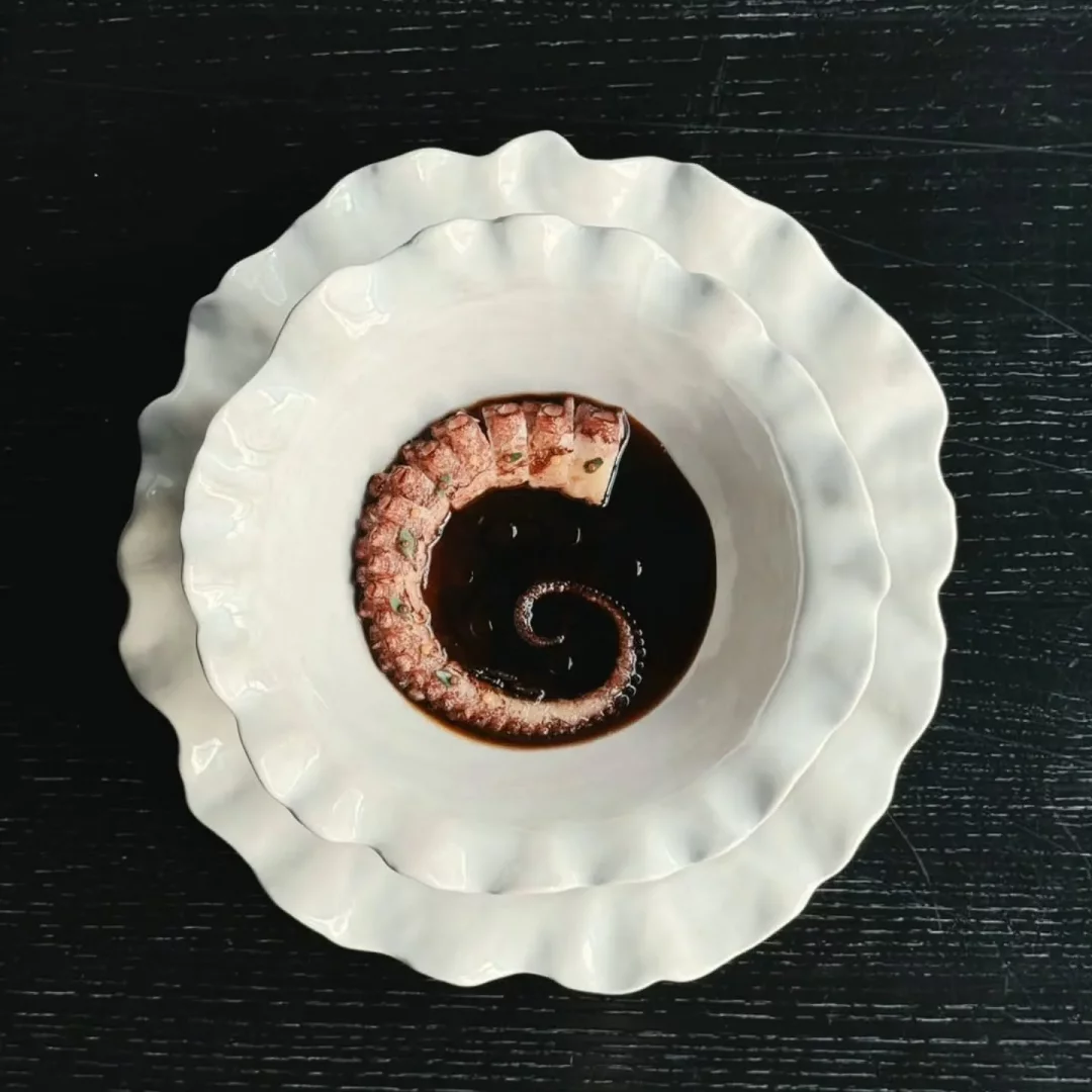 Karanlık ve dokulu bir yüzey üzerinde üst üste duran iki adet beyaz, fırfırlı kenarlı kasenin içinde, koyu renkli parlak bir sosun ortasında zarifçe spiral şeklinde kıvrılmış, dilimlenmiş, kırmızımsı kahverengi bir ahtapot bacağı dikkat çekiyor, üzerindeki küçük yeşil ot parçacıklarıyla modern ve estetik bir yemek sunumu oluşturuyor.