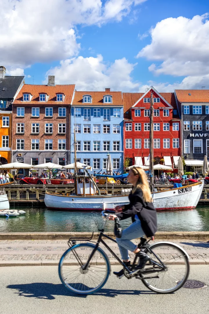 Kopenhag'ın güneşli bir gününde, tarihi Nyhavn bölgesindeki rengarenk cepheli binaların ve kanalda demirli teknelerin oluşturduğu hareketli manzaranın önünden, yolda bisikletiyle hızla ilerleyen sarışın bir kadın şehir yaşamının dinamizmini vurguluyor.