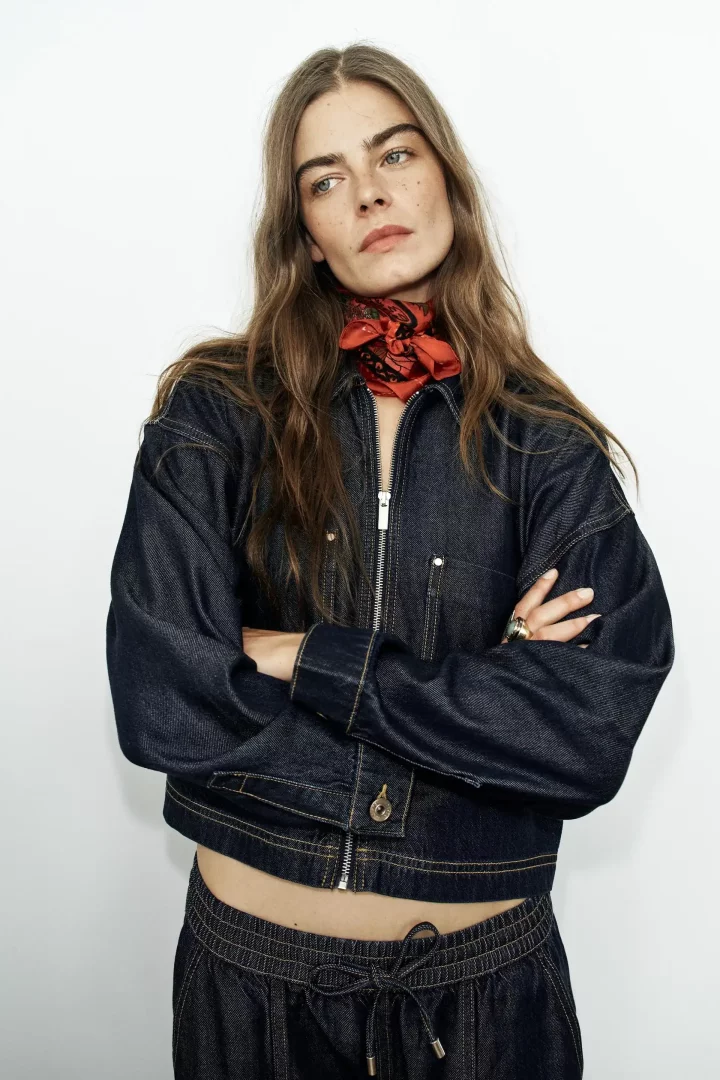 Beyaz fonda, koyu indigo rengi, fermuarlı, kısa kesim bir kot ceket ve paçası lastikli uygun kot pantolon giyen, kolları kavuşmuş, boynunda kırmızı desenli bir bandana olan uzun saçlı genç bir kadın hafifçe sağa bakıyor.