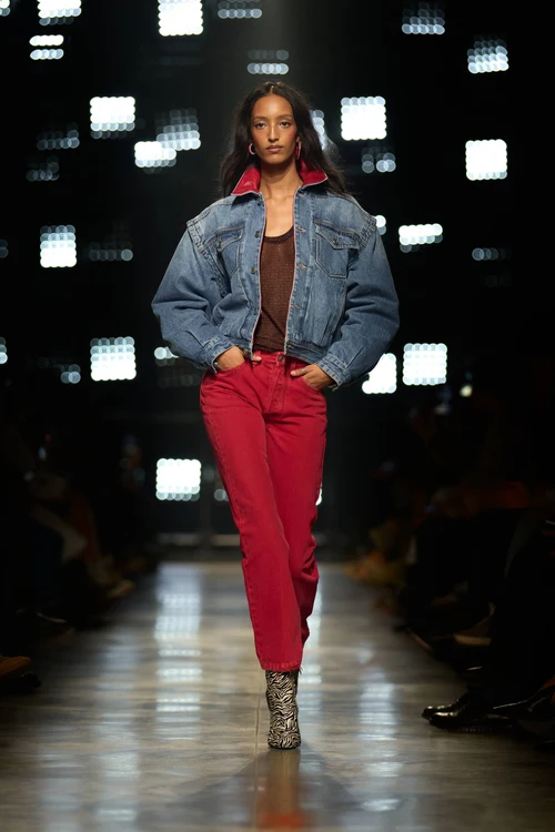 Podyumda yürüyen, kırmızı yakalı bol kesim kot ceket, kahverengi bir bluz, canlı kırmızı pantolon ve zebra desenli bilek botlarla çağdaş bir Isabel Marant görünümü sergileyen model, arkasındaki bulanık ışıklarla dikkat çekiyor.