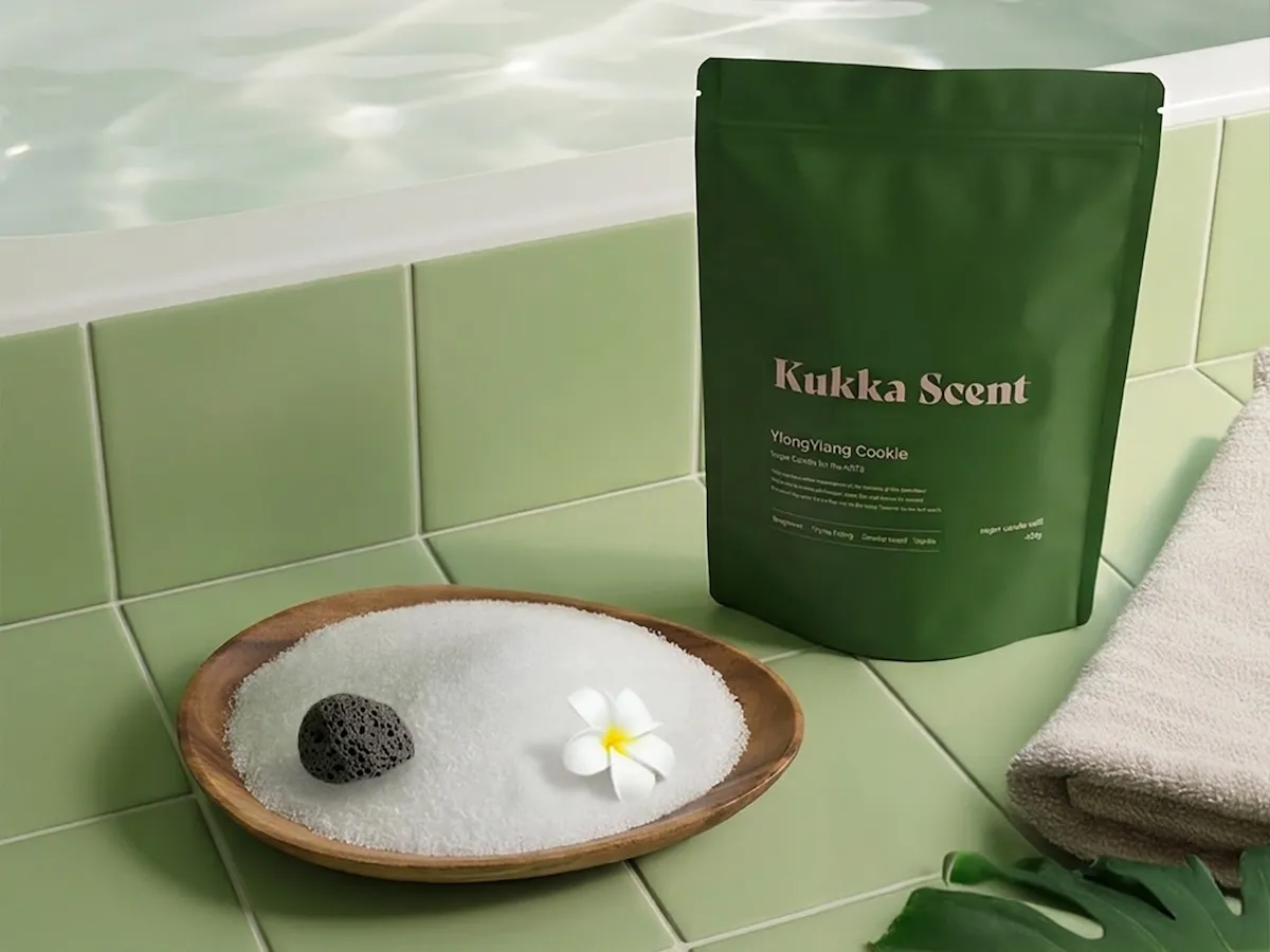 Küvet kenarındaki yeşil fayanslar üzerinde, Kukka Scent markasının YlangYlang Cookie kokulu koyu yeşil banyo tuzu poşeti, beyaz plumeria çiçeği ve ponza taşıyla dolu ahşap bir kase banyo tuzu ile katlanmış krem rengi havlu, dingin bir banyo anı için şık bir düzenleme sunuyor.