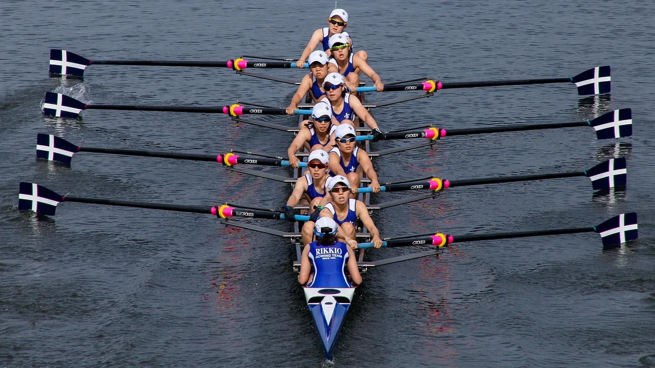 Bir kürek teknesinde sekiz kadın kürekçi ve dümenci, suya batırdıkları beyaz haç desenli küreklerini senkronize bir şekilde çekerek ilerlerken, mavi-beyaz üniformaları ve RIKKIO ROWING TEAM yazısıyla kürek sporunun yoğun rekabet ruhunu yansıtan etkileyici bir görüntü.