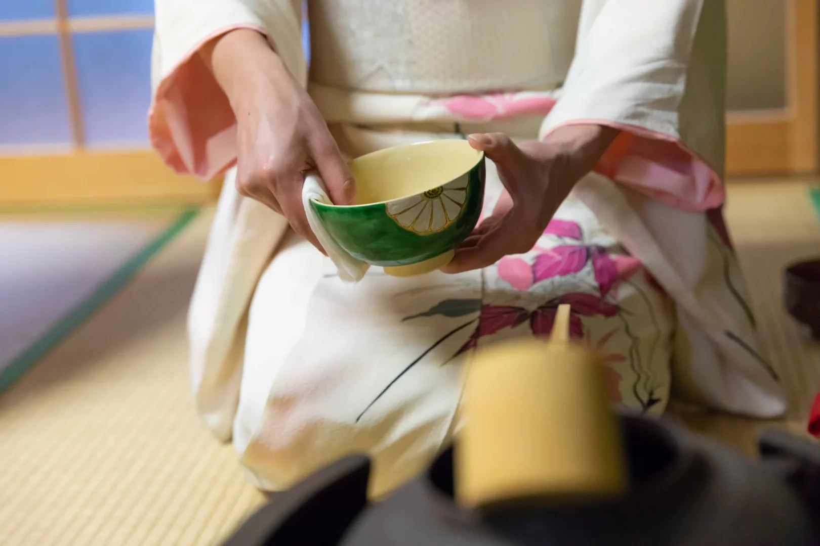 Kyoto'da geleneksel çay seremonisi sırasında, beyaz ve pembe çiçek desenli kimonolu bir kişi tatami minderinde diz çökmüş, zarifçe yeşil ve sarı renklerde desenli bir çay kasesini iki eliyle tutuyor.