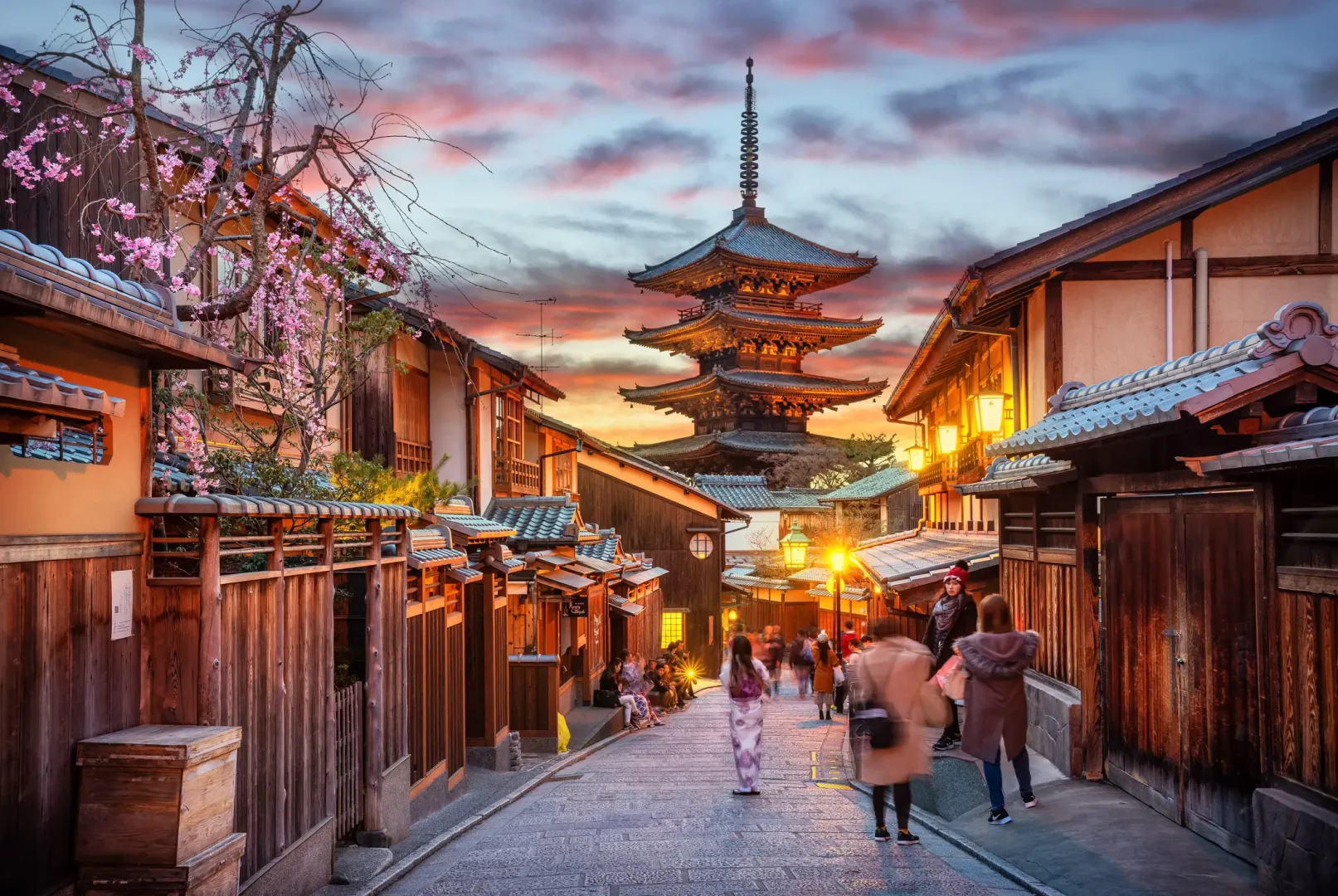 Kyoto'da pembe kiraz çiçekleriyle bezenmiş geleneksel ahşap evlerin ve ışıklı fenerlerin sıralandığı taş döşeli dar bir sokakta, gün batımının kızıl tonlarıyla aydınlanan beş katlı ikonik Yasaka Pagodası'na doğru yürüyen insanlarla dolu, büyüleyici bir manzara.