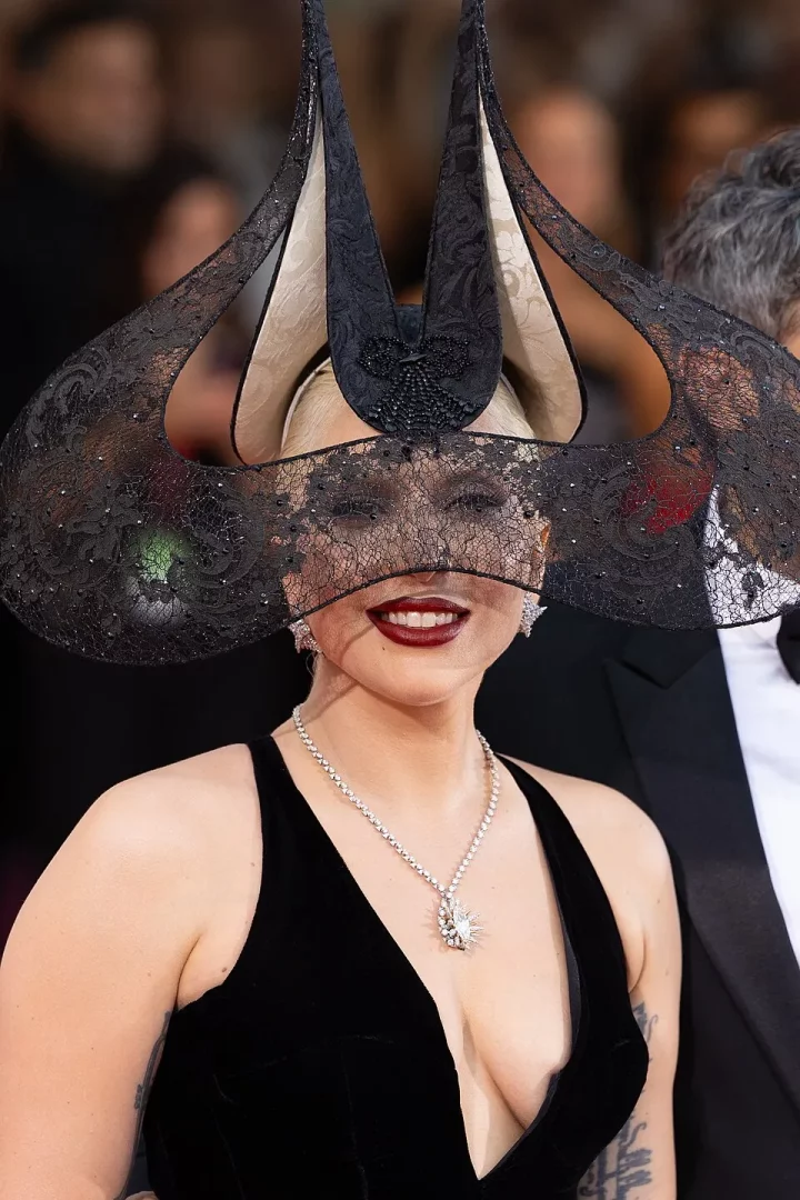 Lady Gaga, kırmızı halıda kendine özgü tarzıyla, yüzünü kısmen kapatan işlemeli siyah dantel peçesi ve sivri uçlu, fütüristik görünümlü büyük şapkasıyla, derin V yakalı siyah kadife elbisesi ve göz kamaştıran elmas kolyesiyle şık bir şekilde gülümsüyor.