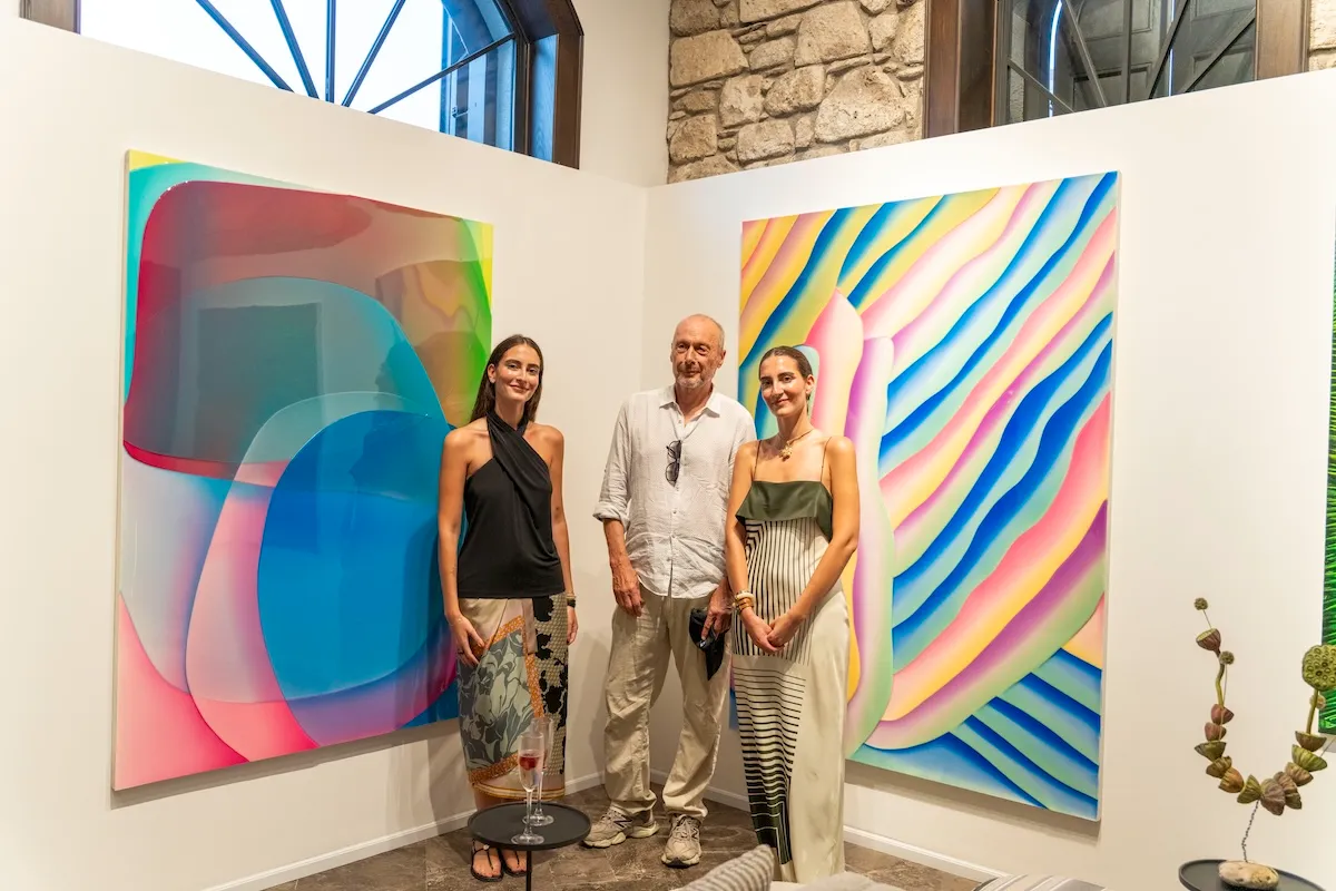 Sanatçı Peter Zimmermann, canlı ve soyut tablolarının sergilendiği bir sanat galerisinde, kucaklayıcı renklerle dolu büyük eserlerinin önünde, Lal Ösen ve Selmin Ösen ile birlikte neşeyle poz veriyor, üçü de samimi bir gülümsemeyle kameraya bakıyor.