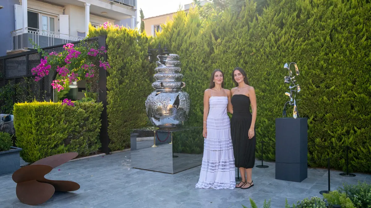 LS Contemporary Heykel Bahçesi'nde, yemyeşil çitler ve mor begonvillerle dolu bahçe zemininde modern bir heykel sergisini gezen ziyaretçiler arasında beyaz ve siyah uzun elbiseleriyle dikkat çeken Lal ve Selmin Ösen, çevredeki çeşitli çağdaş heykellerin önünde gülümsüyorlar.