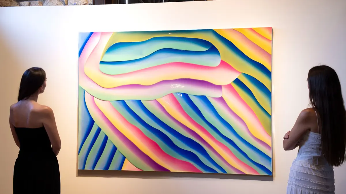 Sanat galerisinde, sırtları dönük iki kadın ziyaretçi, canlı sarı, mavi, pembe, mor ve yeşil tonlarında akışkan, parlak ve kabartmalı soyut dalgalar içeren büyük bir Peter Zimmermann eserini dikkatle inceliyor; Alaçatı'daki bir sergiden bir kare.