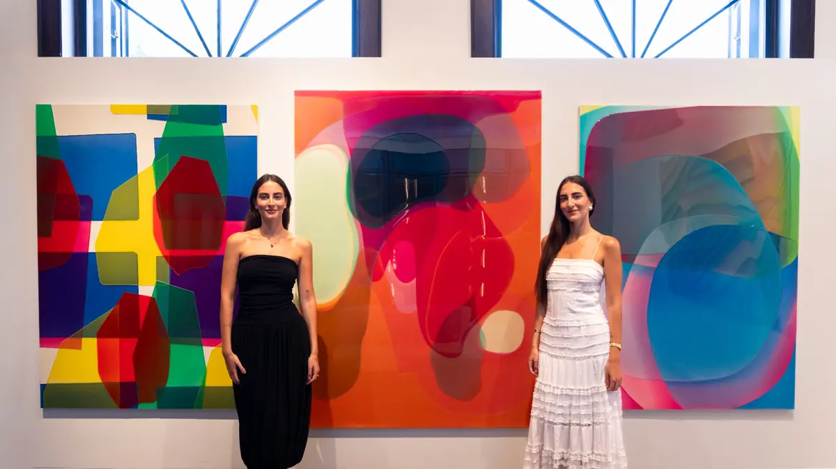 Sanat galerisinde, siyah straplez elbisesiyle Lal ve beyaz askılı, fırfırlı elbisesiyle Selmin Ösen, arkalarında Peter Zimmermann'in canlı renkli, akışkan formlarda katmanlı soyut eserlerinden oluşan üç büyük tablo önünde gülümsüyor.