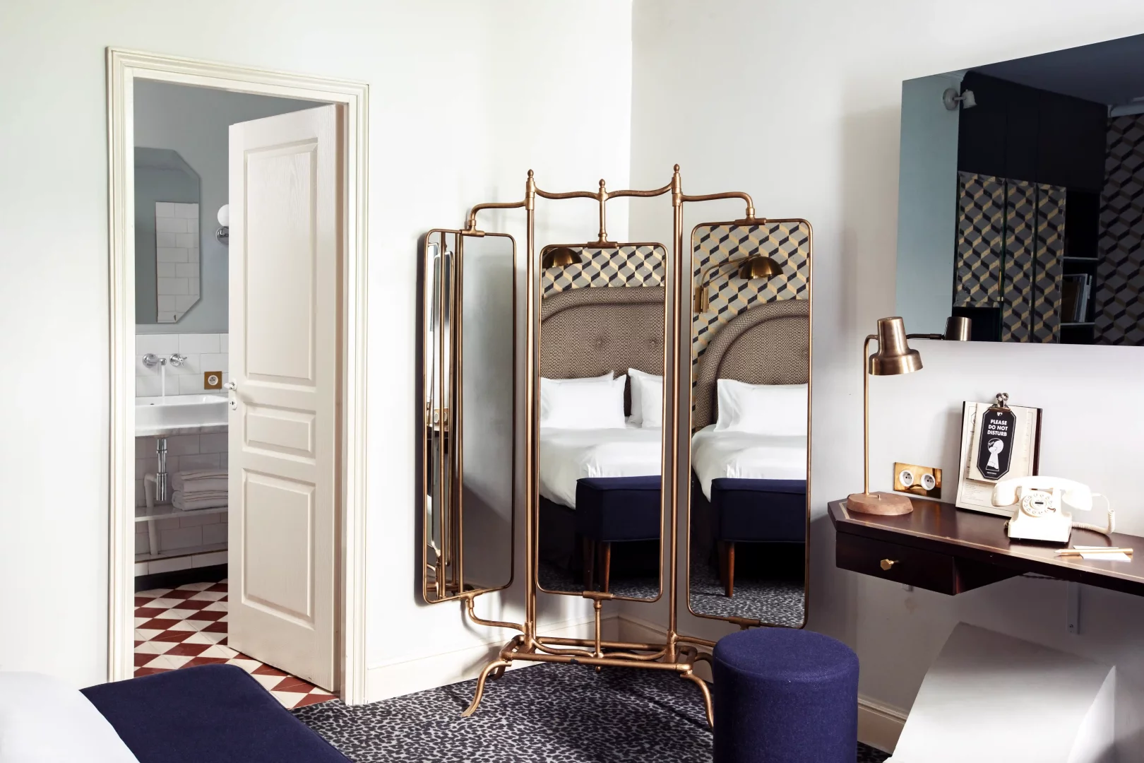 Bu Grand Pigalle Experimental otel odasında, pirinç detaylı aynalar, desenli yatak başlığı ve halı ile şık bir çalışma masası, modern Paris romantizmini yansıtan sıcak ve karakterli bir atmosfer sunuyor.