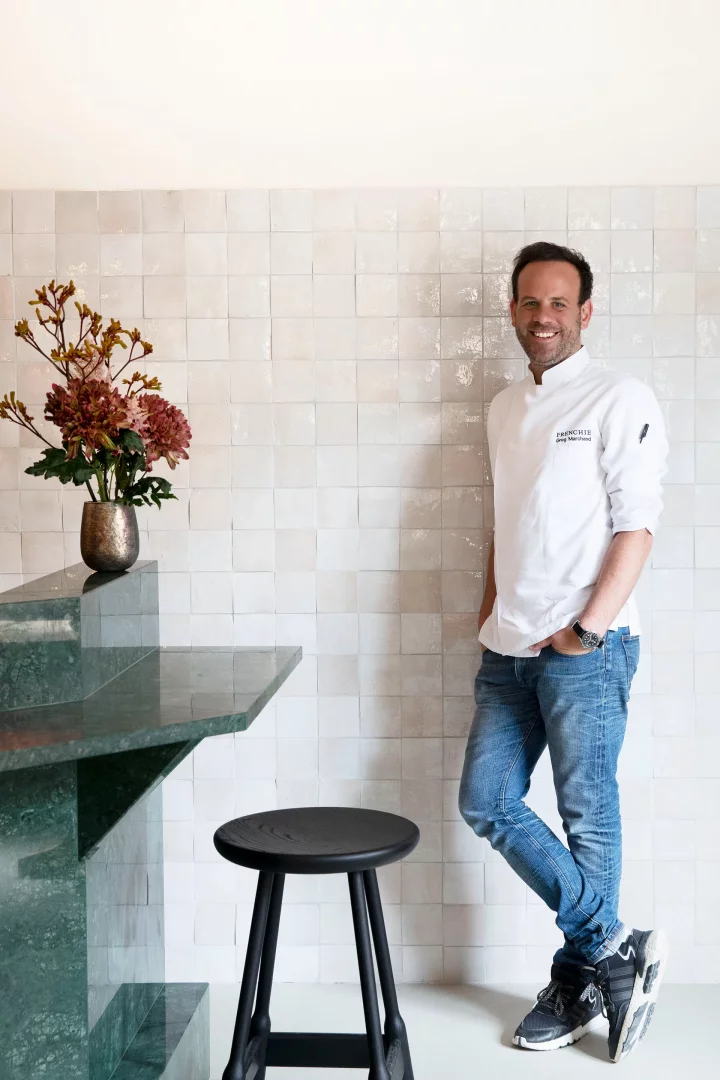 Grand Pigalle Experimental otelinin bünyesindeki Frenchie Pigalle restoranının Michelin yıldızlı şefi Grégory Marchand, çiçeklerle süslü yeşil mermer bir bankoya yaslanmış, beyaz karolarla kaplı duvara karşı gülümseyerek objektife bakıyor.