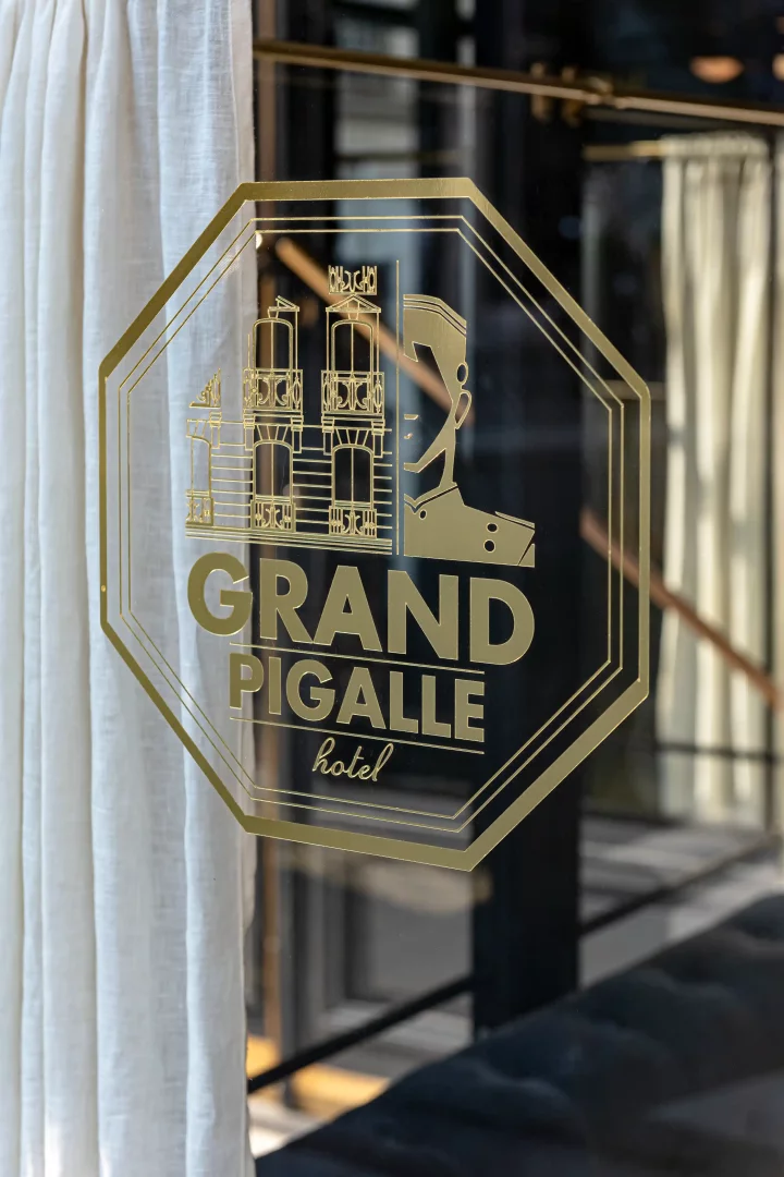 Altın rengi, sekizgen bir çerçeve içinde, üstte stilize bir bina figürü ve altında büyük puntolarla GRAND PIGALLE, daha küçük puntolarla ise hotel yazısı bulunan, cam bir kapı üzerindeki logo.