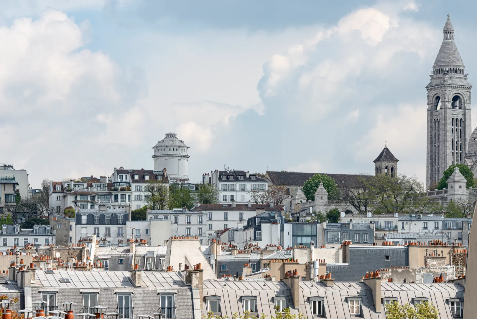 Grand Pigalle Experimental'dan görülen tipik Paris çatıları, bacaları ve bulutlu bir gökyüzü altında Montmartre'ın zirvesinde yükselen, şehrin ikonik simgelerinden Sacré-Cœur Bazilikası'nın ve Montmartre Su Kulesi'nin silüetiyle Paris romantizmini gözler önüne seren büyüleyici bir şehir manzarası.