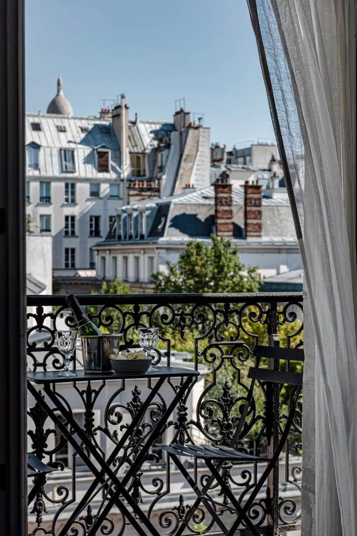 Ferforje balkon korkuluğunun ardında, buz kovasında şampanya, iki kadeh ve bir kase atıştırmalıkla donatılmış siyah bir bistro masası ve sandalyesi, tipik Paris çatıları ve bacalarıyla bezeli güneşli şehir manzarasına bakıyor.