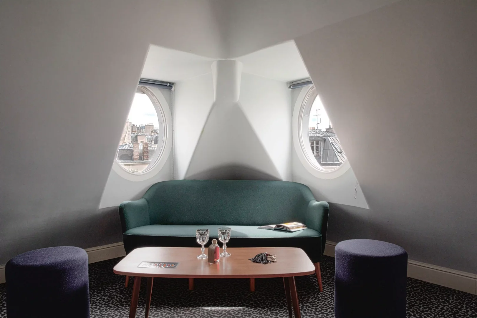 Grand Pigalle Experimental otelinin en üst katındaki bu karakterli odada, oval pencerelerden Paris çatıları manzarasına karşı konumlanmış yeşil kanepe, ahşap sehpa ve mor puflar, Dorothée Meilichzon imzalı modern ve samimi tasarımıyla romantik Paris havasını yansıtıyor.