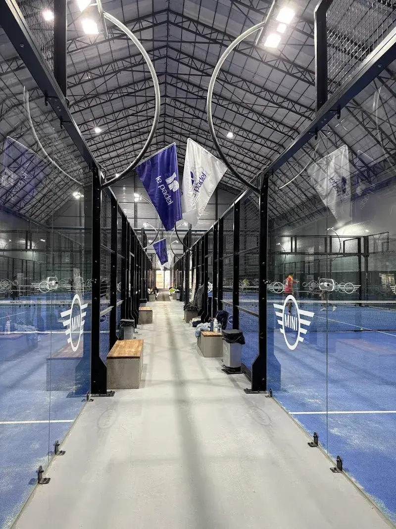 Beşiktaş İstanbul'daki kapalı bir padel tesisinin içinden çekilen bu fotoğrafta, parlak ışıklandırılmış uzun bir koridorun iki yanında, cam paneller ve siyah metal çerçevelerle çevrili mavi padel kortları uzanırken, tavandan sarkan le padel bayrakları ve kortların cam duvarlarındaki MINI logoları modern spor alanını vurguluyor.
