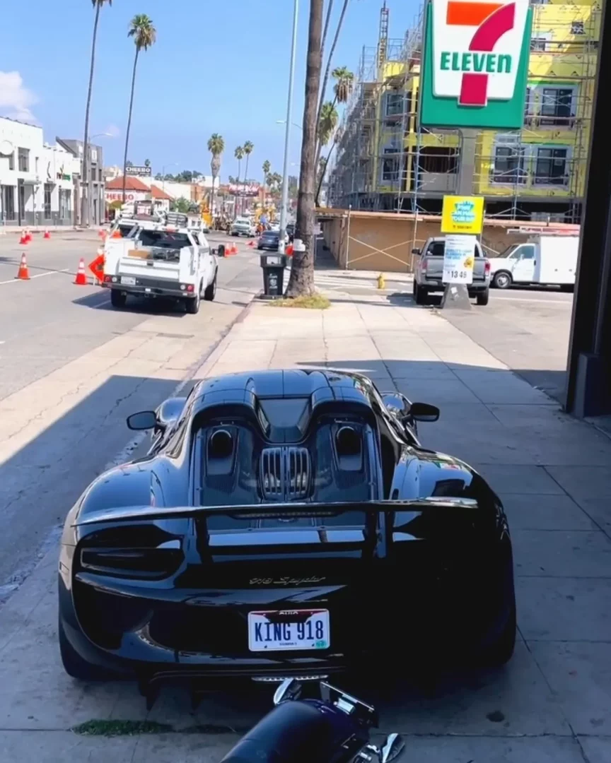 KING 918 plakalı, parlak siyah bir Porsche 918 Spyder'ın arka kısmı, kaldırımda park edilmiş halde görülüyor; aracın güçlü spor tasarımı ve egzoz detayları ön plandayken, arka planda palmiye ağaçlarıyla çevrili bir cadde, yol çalışması konileri ve inşaat halindeki bir binanın üzerindeki 7-Eleven tabelası dikkat çekiyor.