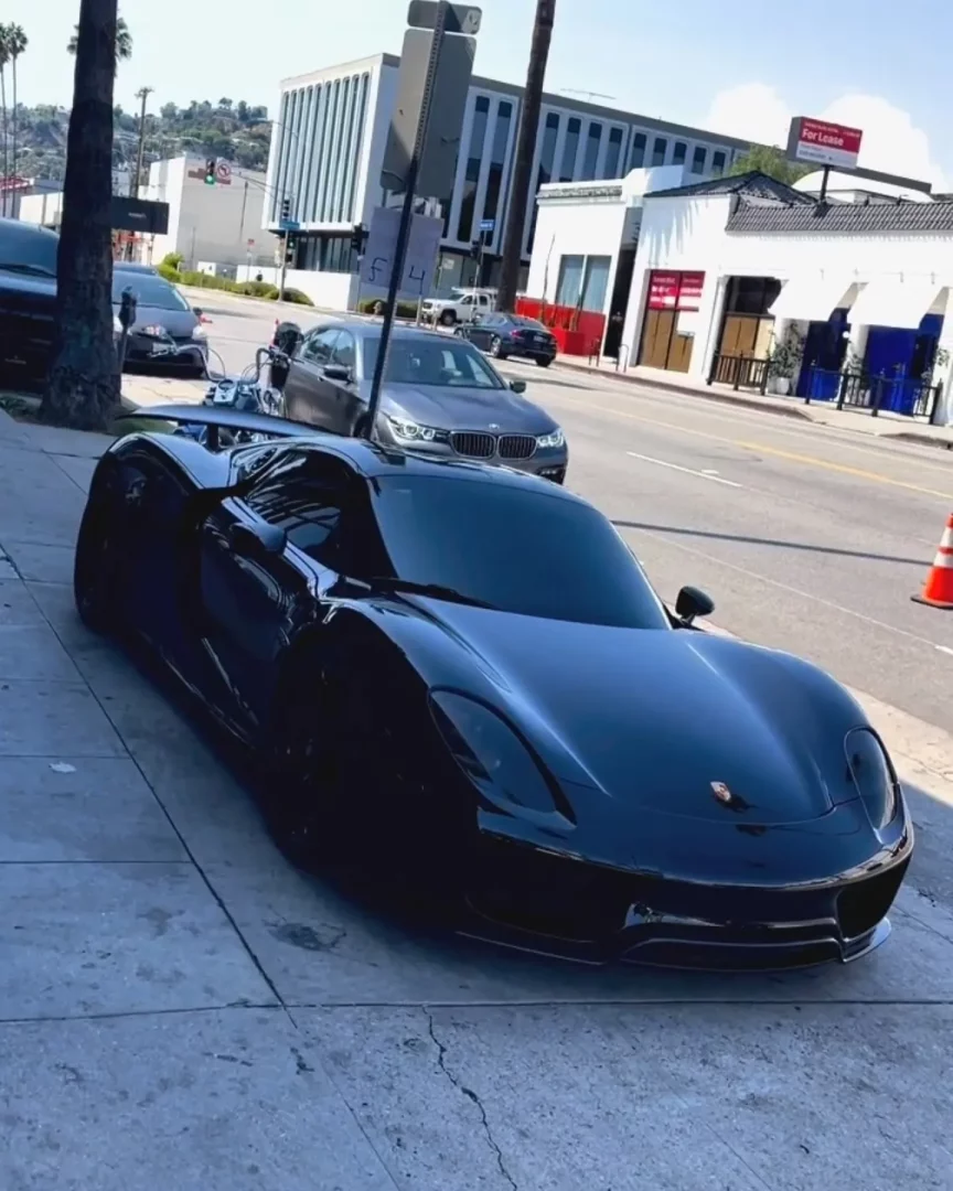 Lebron James'e ait, parlak siyah Porsche 918 Spyder, güneşli bir şehir sokağının kaldırımında park edilmiş durumda; arabanın aerodinamik hatları ve ön kısmındaki Porsche amblemi dikkat çekerken, arka planda palmiye ağaçları ve hareketli bir cadde üzerindeki binalar belirginleşiyor.