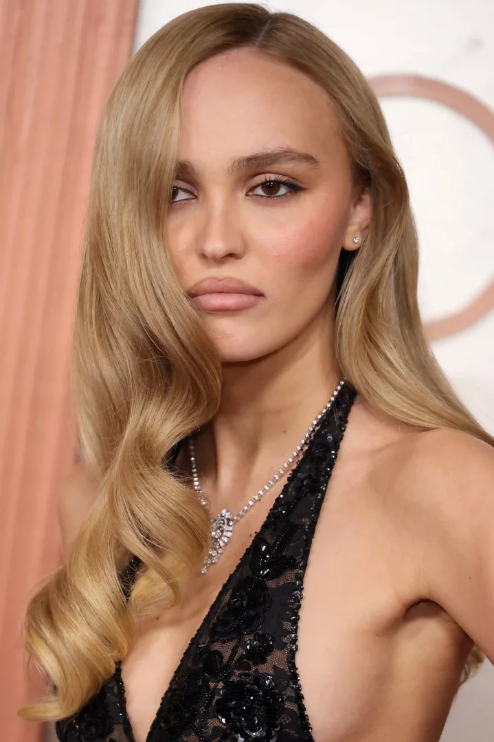 Lily-Rose Depp, yana ayrılmış, yüzünü çerçeveleyen ve omzuna dökülen belirgin iri dalgalara sahip sarı saç modeliyle dikkat çekerken, derin V yakalı siyah dantel ve payet işlemeli üstü ile ışıltılı bir kolye takıyor.