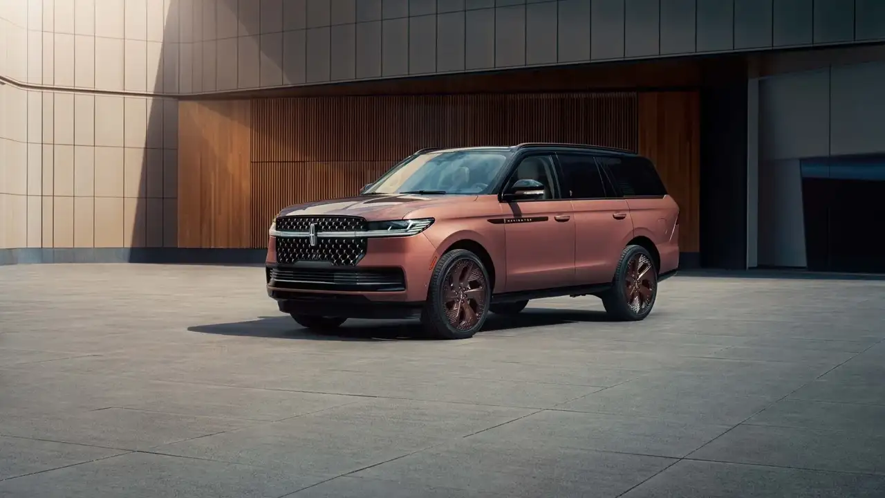 Modern mimariye sahip bir binanın önündeki geniş avluda, rose gold rengi, siyah tavanı ve bronz jantlarıyla dikkat çeken, zarif ve özel tasarım bir Lincoln Navigator SUV aracı park edilmiş halde duruyor.
